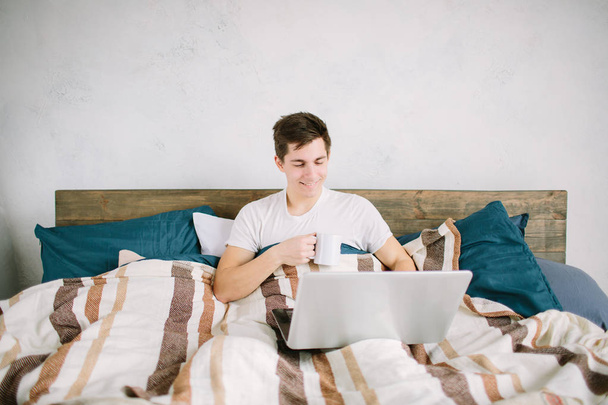 casual young man using laptop in bed at home and drinking coffee - Foto, immagini