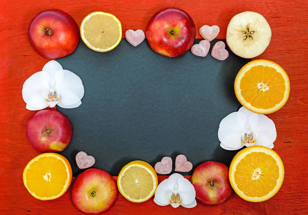 Bright multicolored background (frame) with citrus (lemon, orange), cutting the apples, jelly sweets in the shape of a heart, white  flower orchid on a red board. In the middle of the black rectangle - empty space for your projects, notes. - Foto, Imagem