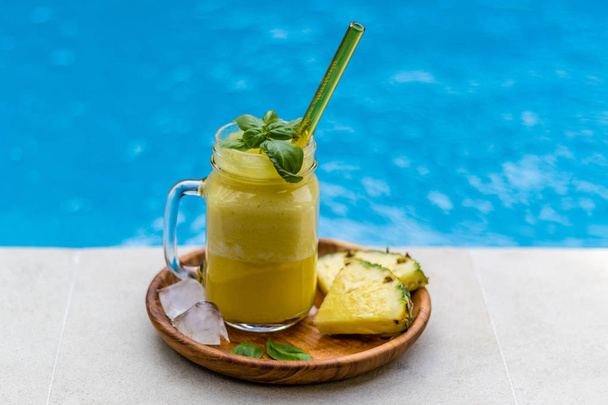 Lasi tuoretta ananasmehua keltaisella lasipillillä, tummalla puulevyllä, tarjoillaan kahden ananaspalan, jääkuutioiden ja basilikan lehtien kanssa uima-altaan reunalla. Sivukuva
 - Valokuva, kuva