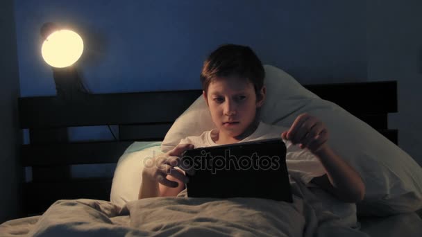 Boy resting in his bed in the evening using his digital tablet turning off light and falling asleep - Кадры, видео
