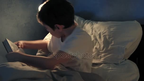 Boy resting in his bed in the evening and reading a book - Πλάνα, βίντεο