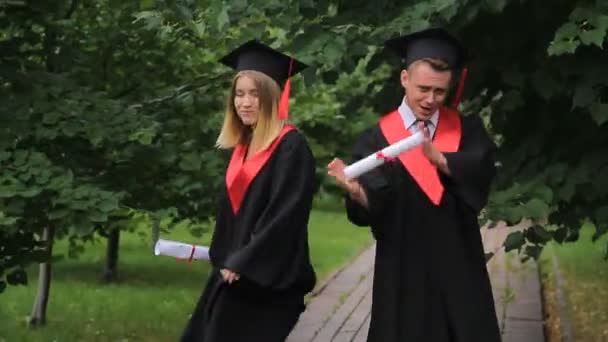 Szczęśliwi młodzi absolwenci tańczą i świętują ukończenie szkoły w parku niedaleko akademii - Materiał filmowy, wideo