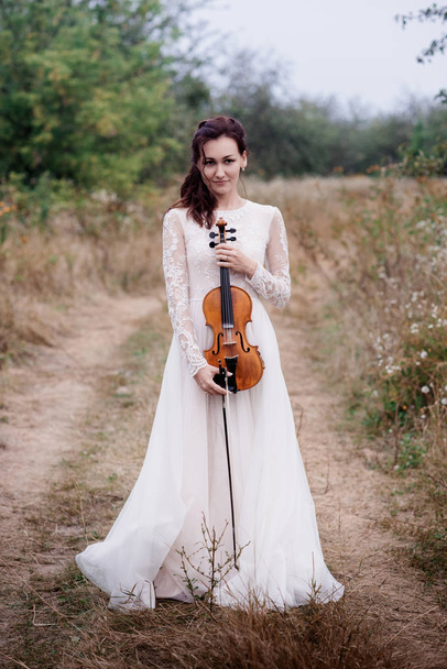Young beautiful girl in a white dress under tree holding a violin in hands. - Фото, зображення