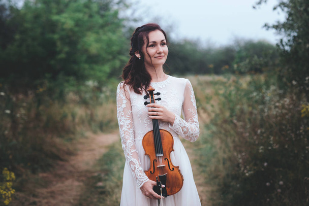 Молода красива дівчина в білій сукні під деревом тримає скрипку в руках
. - Фото, зображення