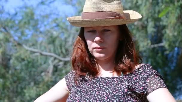 junge schöne Frau mit roten Haaren und Strohhut, die draußen sitzt und in die Kamera schaut und dann ernst aufblickt - Filmmaterial, Video