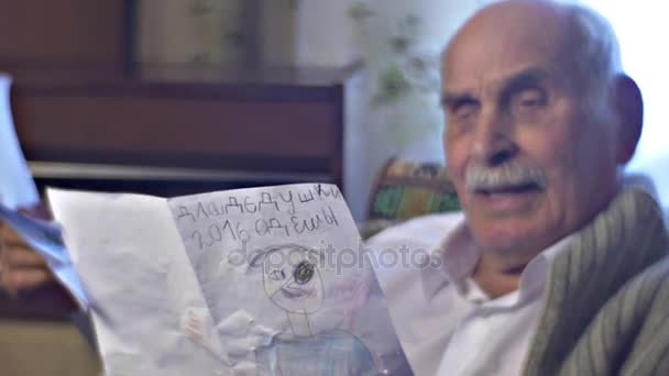 Senior Man is Glad to Receive a Gift From Kids Holding the Drawings and Shows it Proud of His Happy United Family Grandad is Not Lonely in Cosy Room - Πλάνα, βίντεο