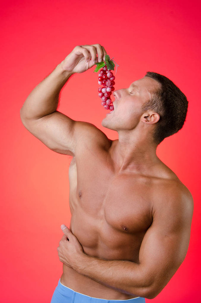 Σέξι άνδρας τρώγοντας σταφύλια. Κόκκινο φόντο. - Φωτογραφία, εικόνα