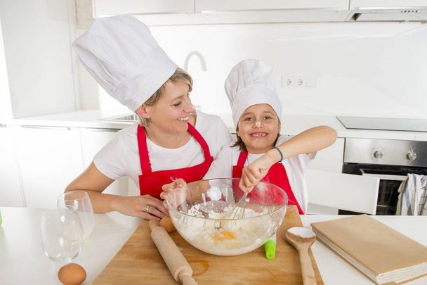young mother and little sweet daughter in cook hat and apron cooking together baking at home kitchen - Photo, Image