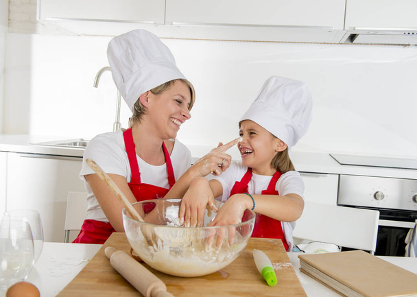 νεαρή μητέρα και κορούλα γλυκό σε μάγειρας καπέλο και ποδιά μαγειρικής μαζί ψήσιμο στο σπίτι κουζίνα - Φωτογραφία, εικόνα