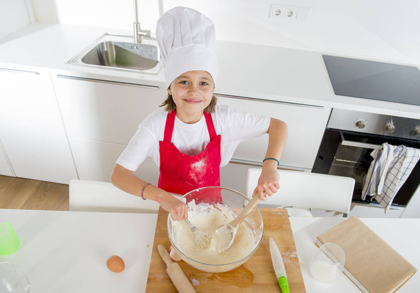 mini chef chica con sombrero de cocinero y delantal mezcla de harina y huevos hornear preparación dulce desierto sonriendo feliz
 - Foto, Imagen