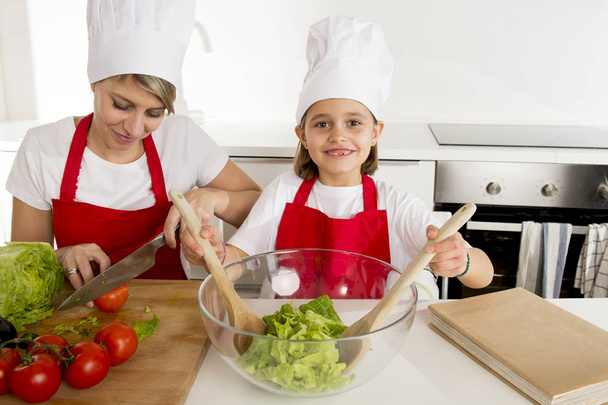 mother and little daughter cooking together with hat apron preparing salad at home kitchen - Fotó, kép