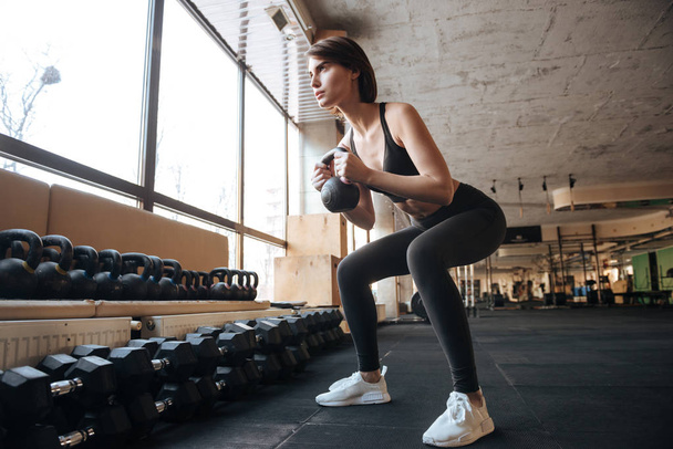 Sportswoman working our and doing squats with kettlebell in gym - Fotoğraf, Görsel