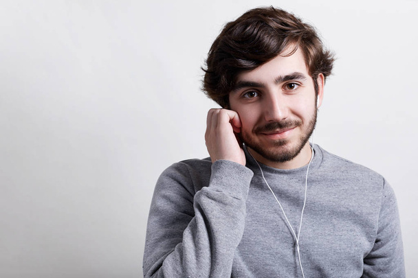 Young hipster with big dark eyes modern hairstyle and beard wearing casual grey sweater listening to the music or audiobook with his earphones holding his hand on earphones having happy expression - Photo, Image