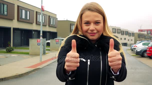 A young, beautiful woman stands on the street in a suburban area and shows thumbs up to the camera with a smile - Footage, Video