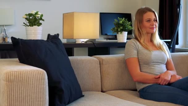 Μια νεαρή, όμορφη γυναίκα κάθεται σε έναν καναπέ στο σαλόνι και ρολόγια τηλεόραση που είναι από την κάμερα - Πλάνα, βίντεο
