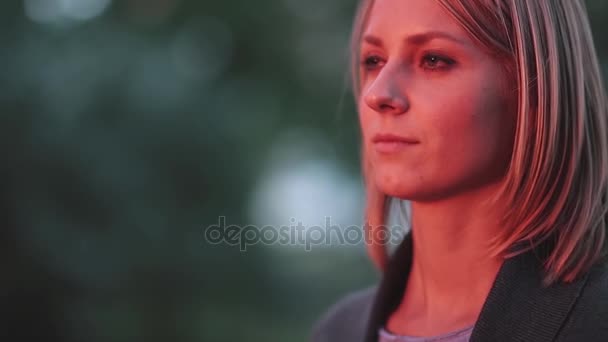 Attractive young woman standing at park in the rays of the setting sun and looking into the distance. Close-up - Filmati, video