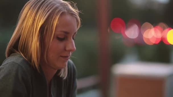 Side view of beautiful young woman sitting in cafe and looking into the distance. Close-up, blurred lights, urban street - Video