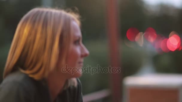 Attractive smiling young woman sitting in the city cafe. Close-up, blurred lights, urban street, side view - Кадри, відео