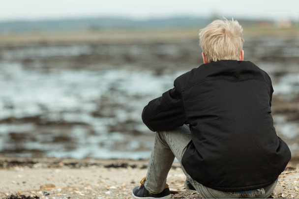 Вид сзади на блондинку, сидящую на скалистом пляже
 - Фото, изображение