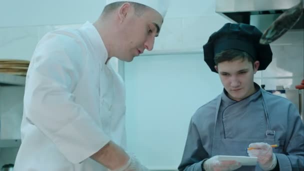 Chef instruyendo a aprendiz en la cocina mientras está tomando notas
 - Metraje, vídeo