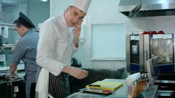 Koch telefoniert und wirft in der Restaurantküche Zutaten auf eine Pfanne - Filmmaterial, Video