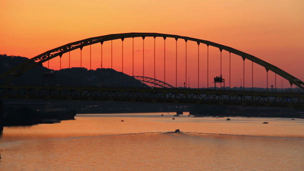 El tráfico pasa sobre el puente de Fort Pitt durante el atardecer en Pittsburgh, PA como barcos de recreo viajan debajo
. - Imágenes, Vídeo