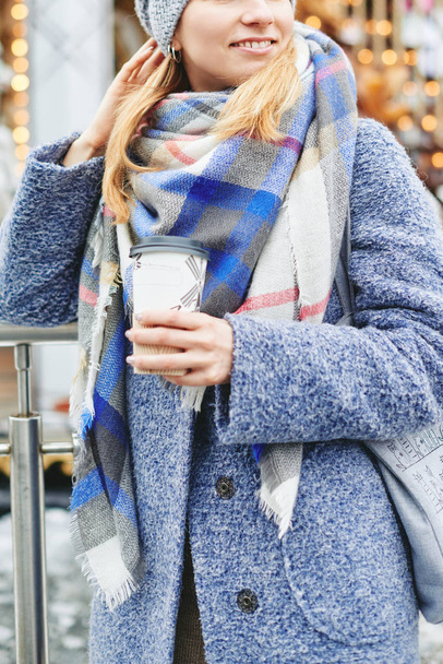 Girl in grey coat, hat and blanket scarf with cup of coffee near carousel - Photo, image