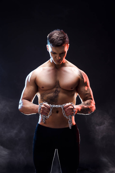 Handsome power athletic man bodybuilder doing exercises with chain, tearing. Fitness muscular body on dark background. Perfect male. Awesome bodybuilder, tattoo, posing. - Fotó, kép