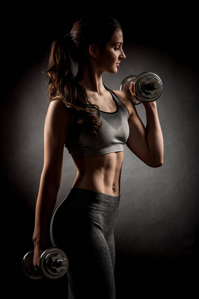 Atractive fit woman works out with dumbbells as a fitness conceptual over dark background. - Foto, imagen