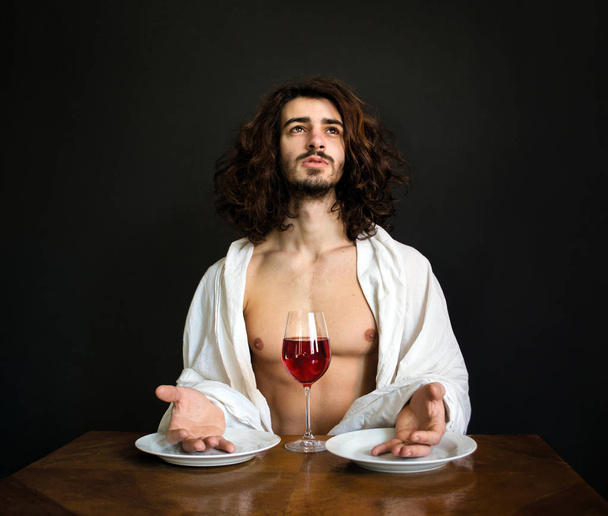 fotó félig meztelen jóképű srác az asztalnál, a két fehér üres lemez hosszú göndör haja, és imádkozik egy pohár vörös bor - Fotó, kép