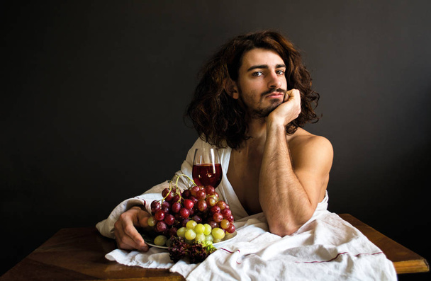 Foto van de knappe jongen met lang krullend haar op de tabel met glas rode drankje en een plaat van druiven - Foto, afbeelding