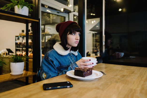 Young hipster girl eats tasty chocolate cake and drinking hot tea in a beautiful cafe - Photo, Image