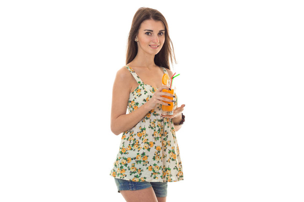 beauty young girl in jeans shorts and sarafan with floral pattern drinks orange cocktail and looking at the camera isolated on white background - Foto, Imagem