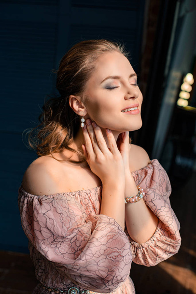 beautiful young blond woman on black background, rich glamorous woman in a pink evening dress with bare ssoulders, the happy woman the sun shines from the window - Foto, Bild