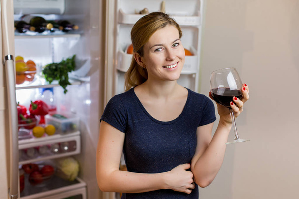 Mujer joven en ropa de dormir bebiendo vino tinto cerca del refrigerador
 - Foto, imagen
