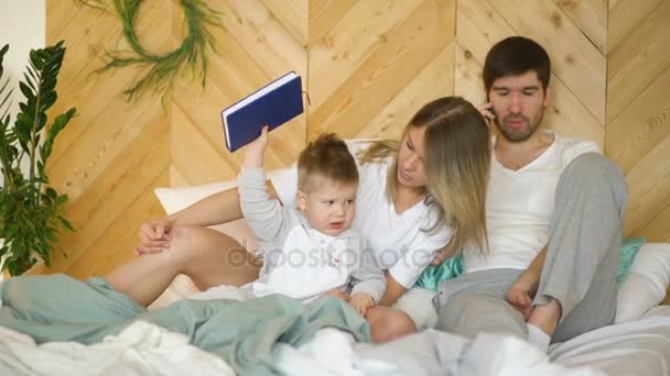 Young mother plays with son while father talking phone on bed - Séquence, vidéo