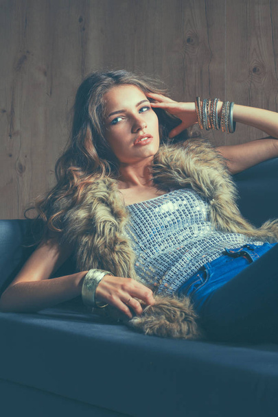 Portrait of elegant woman sitting on black sofa wearing a blue jeans and fur vest - Fotografie, Obrázek