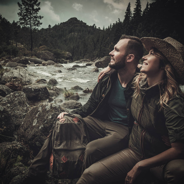 Cheerful couple hikers sitting near wild mountain river. - Zdjęcie, obraz