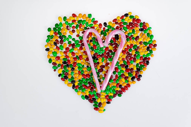 Coloured jelly beans are scattered in the shape of heart and sweet pink and white striped cane on a white background. - Foto, afbeelding