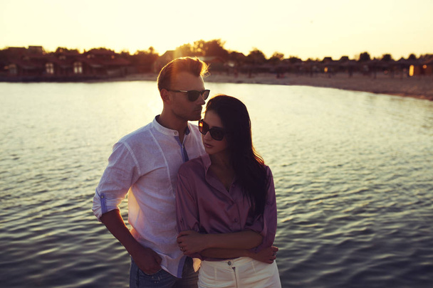 Молодая счастливая модная пара отдыхает у моря на закате. Мужчина и женщина в романтической сексуальной летней одежде и в стильных модных солнечных очках
 - Фото, изображение