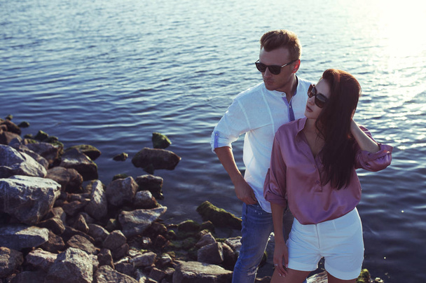 Mladí módní snoubence odpočívá u moře na západ slunce. Muž a žena na sobě v romantických sexy letní oblečení a stylové módní sluneční brýle - Fotografie, Obrázek