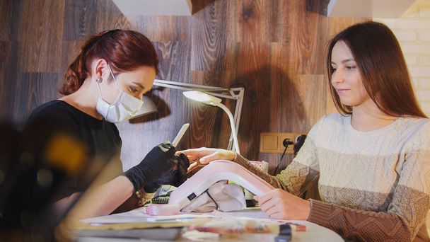 Manicurist in medical mask doing manicure for attractive woman in beauty salon - Photo, Image