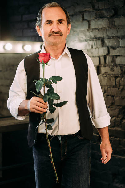 中年男性は白いシャツと黒いベストに身を包んだ赤いバラの花を保持しているハンサムな中間独身。黒の背景. - 写真・画像