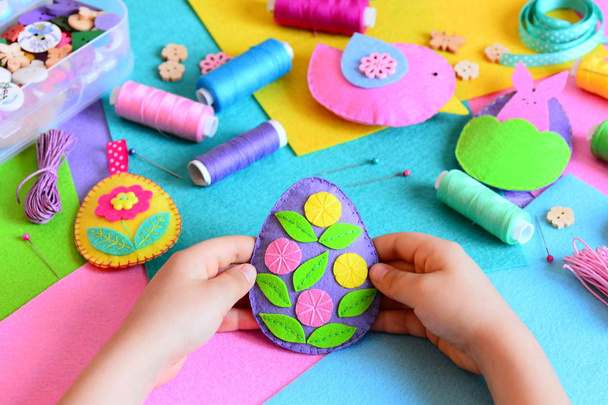 Child made Easter egg decor from felt. Small child holds a felt Easter egg decor in his hands. Easter crafts set, handicraft tools and materials on a table. Holiday spring crafts concept  - Photo, Image