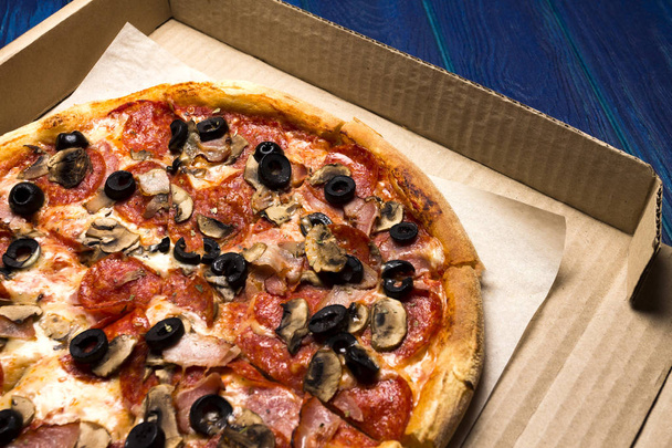 pizza with pepperoni, olives, bacon, mushroom, cheese in the pizza box (corrugated cardboard) on the wooden background - Foto, Imagem