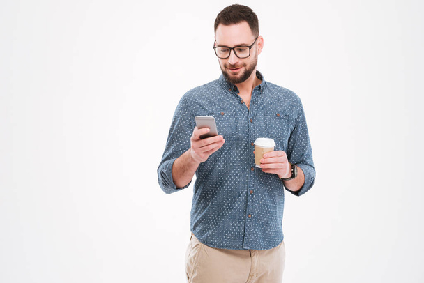 Cheerful bearded man standing with cup of coffee and smartphone - Photo, Image