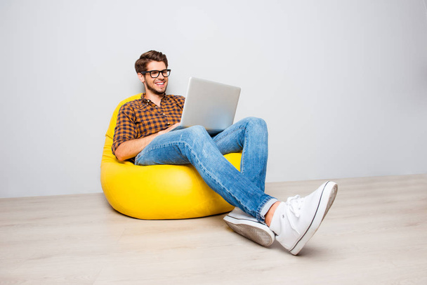 Happy young man sitting in yellow pouf  and using laptop - 写真・画像