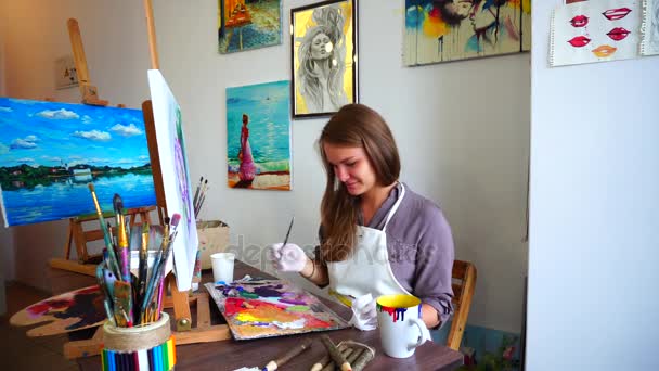 Κορίτσι καλλιτέχνης χρώματα σε καμβά. Ζωγράφος αναμιγνύει χρώμα στην παλέτα και βούρτσα απεικονίζει λουλούδια που κάθεται σε καρέκλα στο τραπέζι στην τάξη τέχνη. - Πλάνα, βίντεο