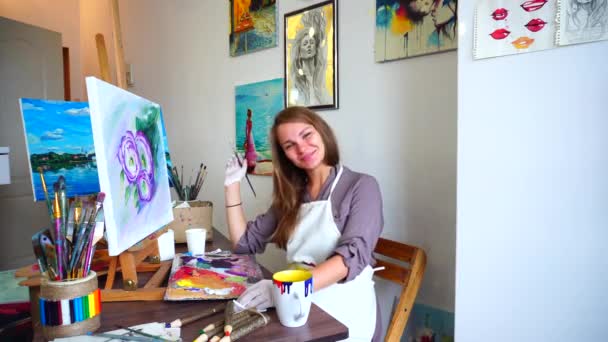 gut aussehende Künstlerin blickt in die Kamera und lächelt, während sie Pinsel zum Malen hält und sitzt am Tisch in einem geräumigen Kunstatelier. - Filmmaterial, Video