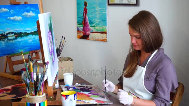 Portrét umělce na stojan. Dívka se mísí barvy na paletě s štětec a barvy na plátně obrázek, sedí na židli u stolu ve výtvarce. - Záběry, video